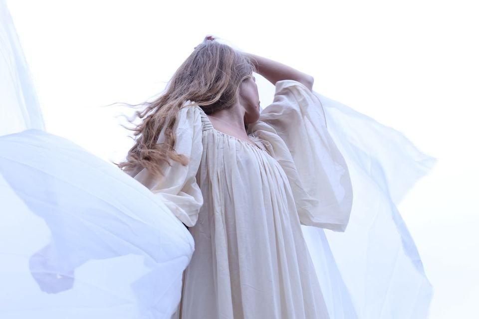 Seen from below against a white sky, a woman in a cream dress raises her arm as a sheer veil billows around her.