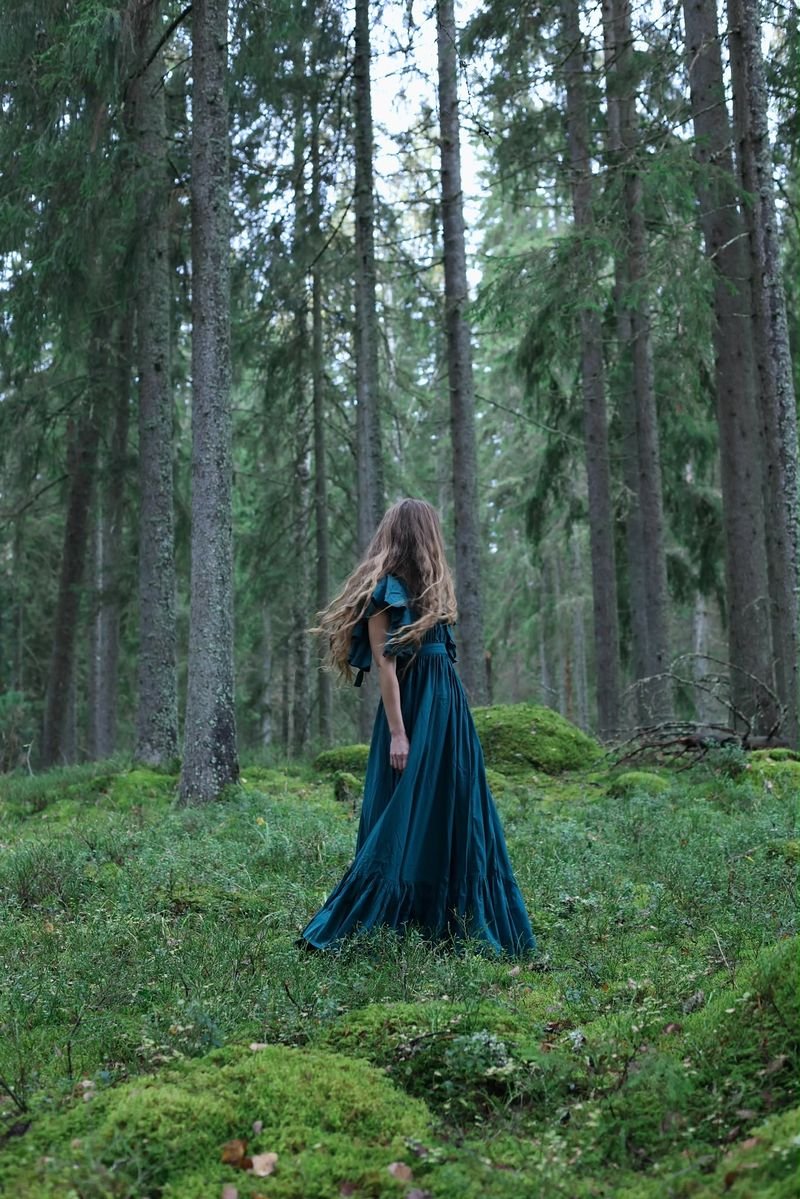 A woman in a long teal dress turns among tall spruce trees on a mossy forest floor, her hair in motion.