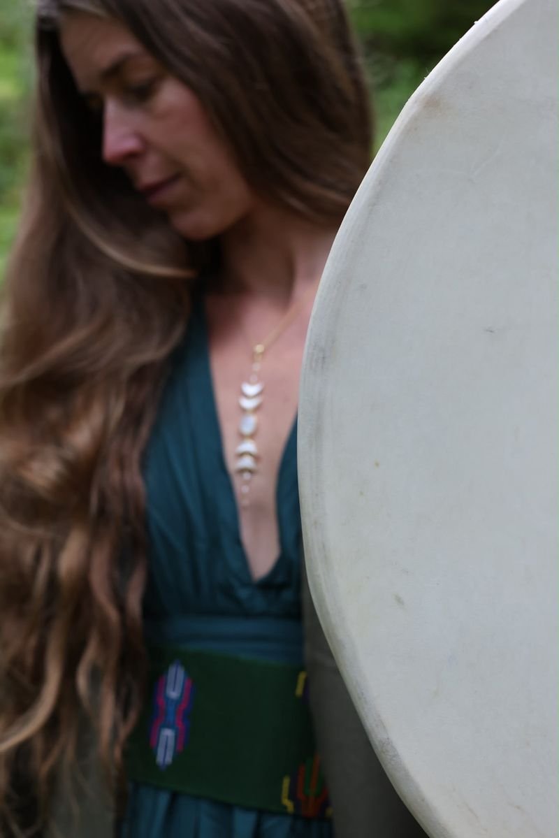 A woman with long hair looks down beside a large frame drum, wearing a deep green dress, a woven belt and a long pendant necklace.
