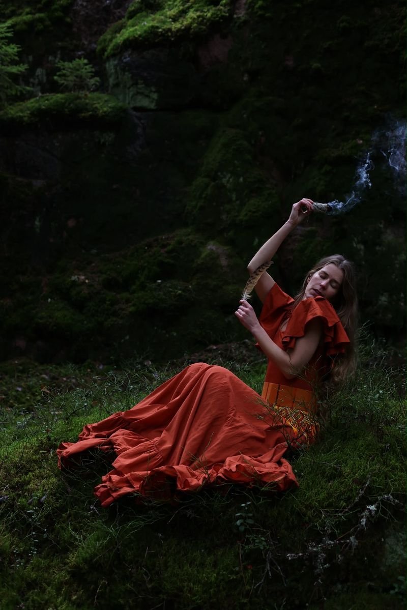 A woman in a rust-red dress sits on the moss of a dark forest, raising a smoking bundle of herbs and holding a feather.