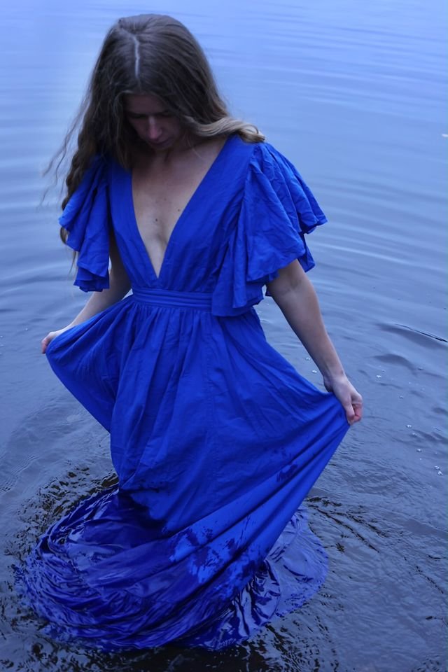 A woman in a cobalt-blue dress wades into still water, holding her skirt as it spreads on the surface.